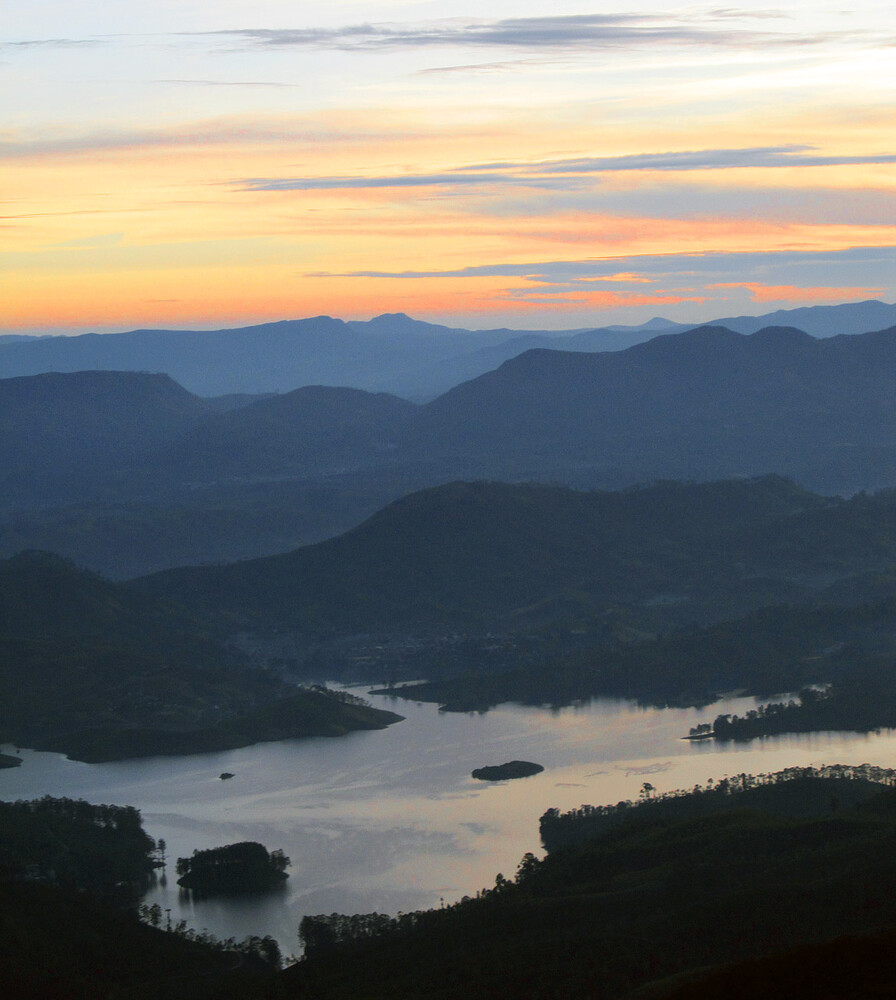 Layered blue mountain ridges fading into haze under a pastel orange and purple sunrise sky, with a calm lake and wooded islands below.
