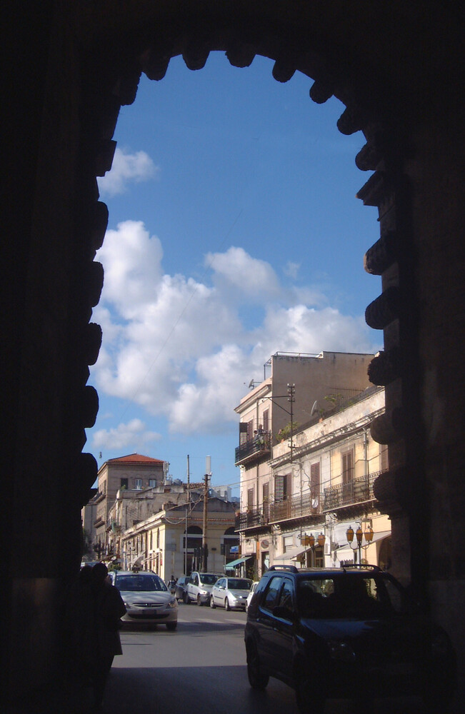 Framed view through a dark stone archway of a sunlit street with parked cars and pastel-facade buildings under a blue sky with clouds.