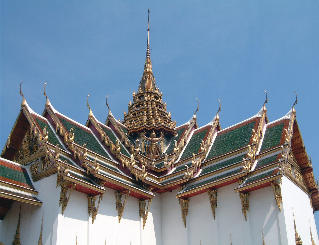 Low-angle view of a Thai temple roofline with tiered green and red tiles, gilded ornaments, and a golden spire against a clear blue sky.