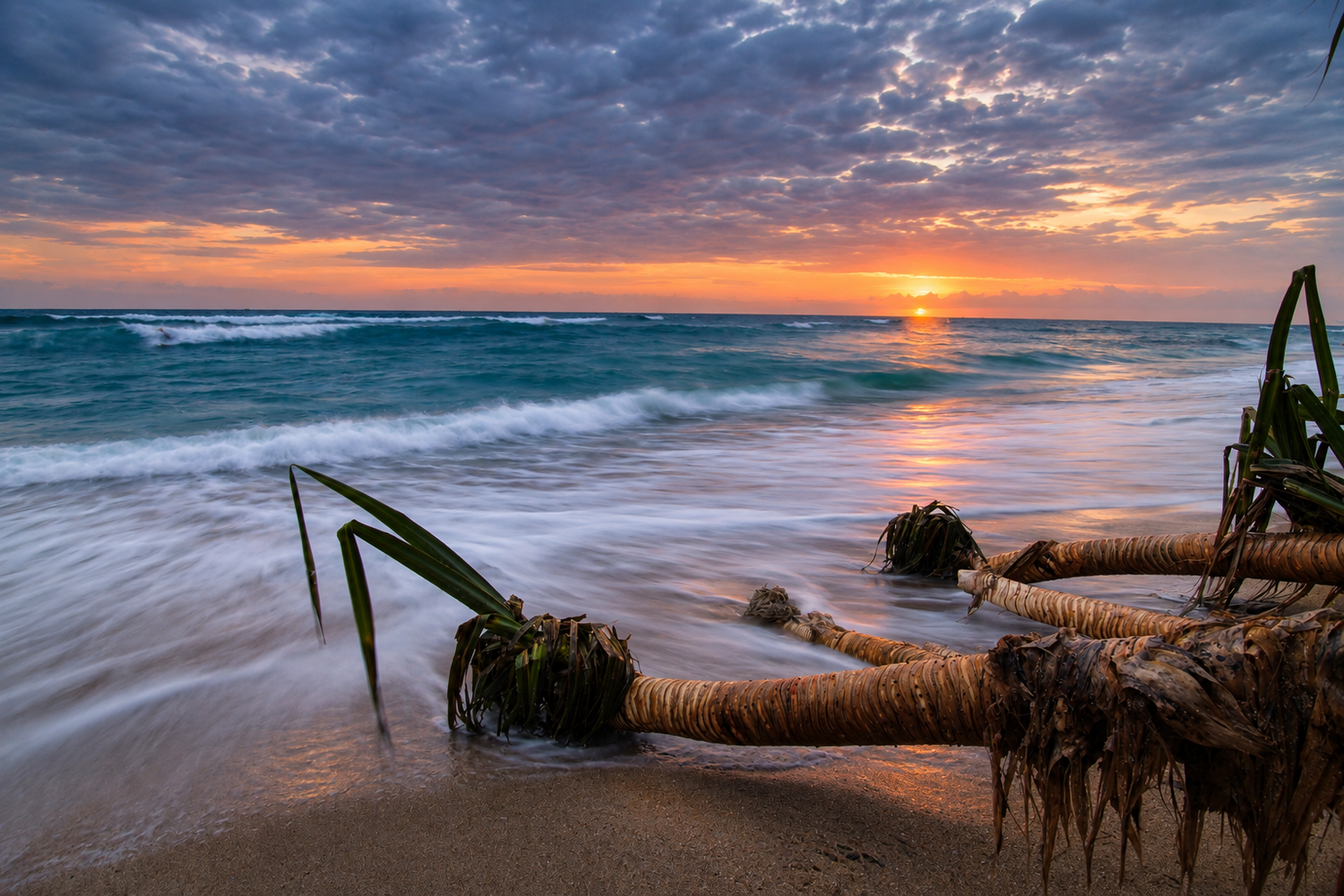 Weathered wooden posts emerging from shallow waves at sunrise, with dramatic cloud-streaked sky