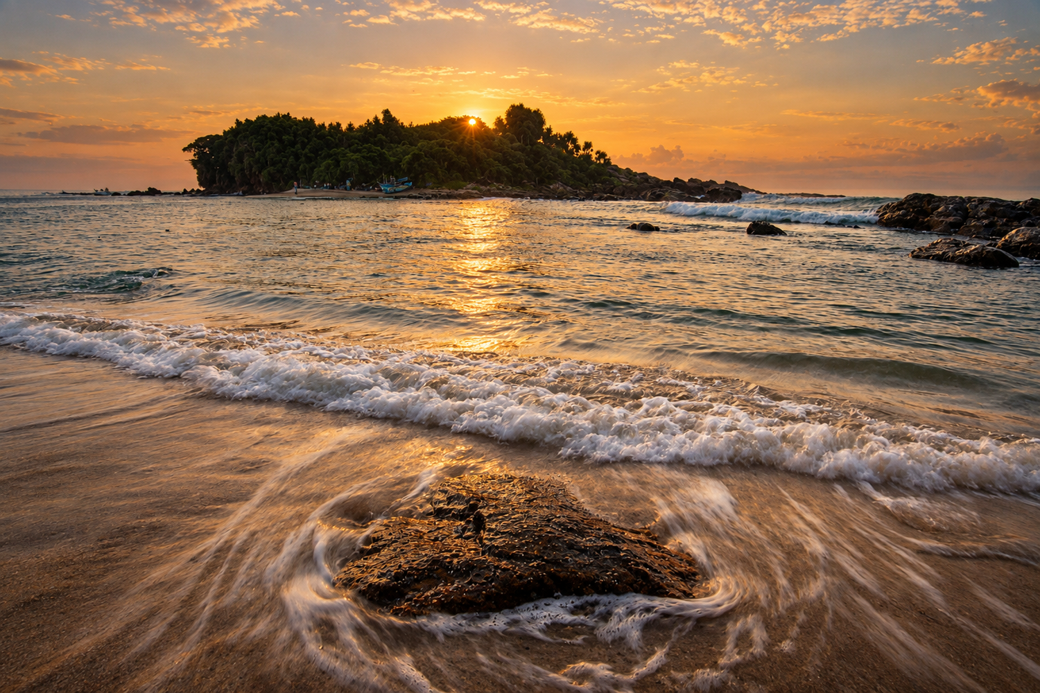 Coastal beach at sunset with waves, rocky foreground, and forested island silhouetted against golden sky