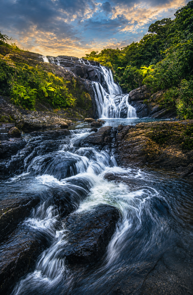 Waterfall cascading over dark rocks with long exposure blur effect and forested hillside at sunset