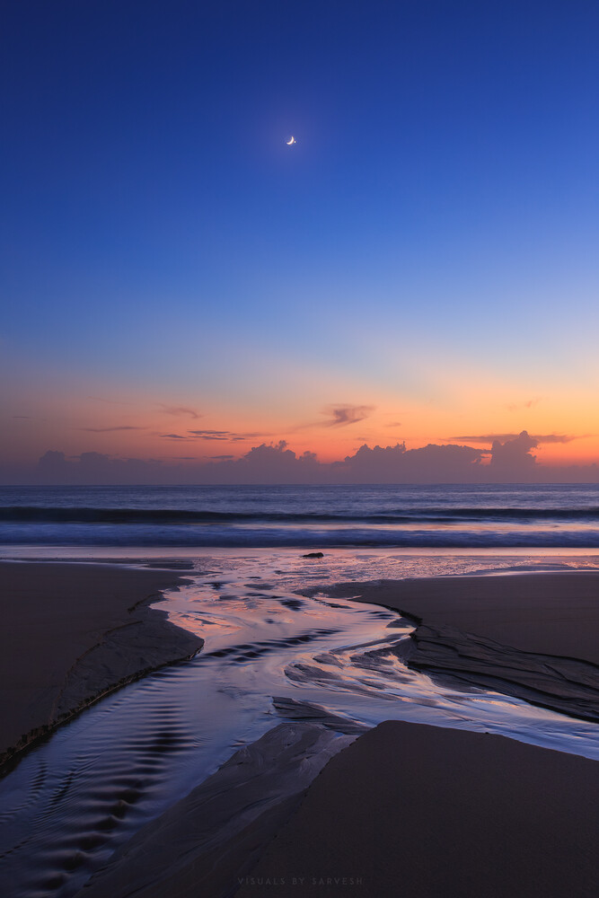 Twilight beach landscape with tidal channels reflecting sunset colors and a crescent moon