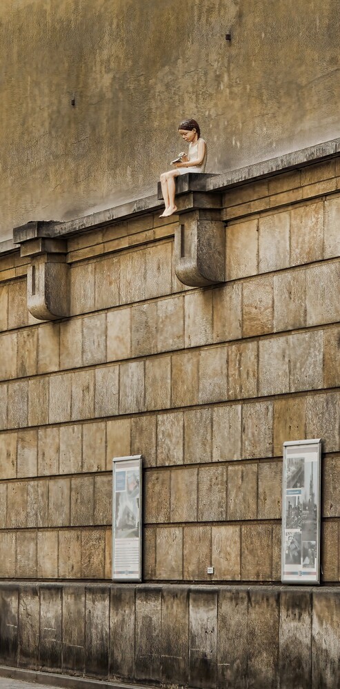Small figure sitting on the top ledge of a tall tan stone wall, with a textured paneled facade and two plaques below.