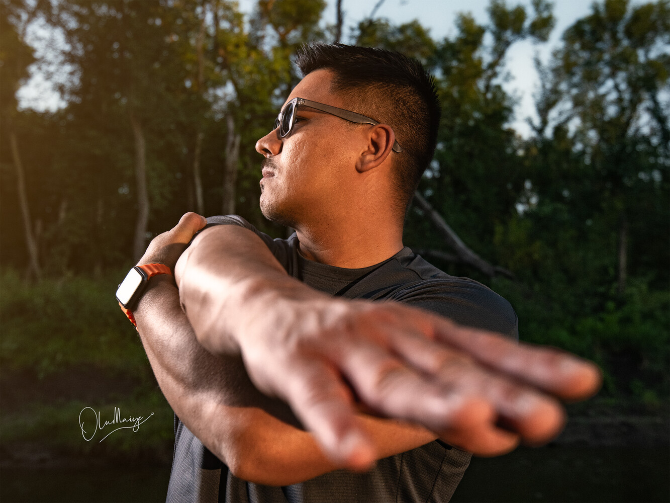 Man in glasses and dark shirt extending arm toward camera in outdoor forest setting