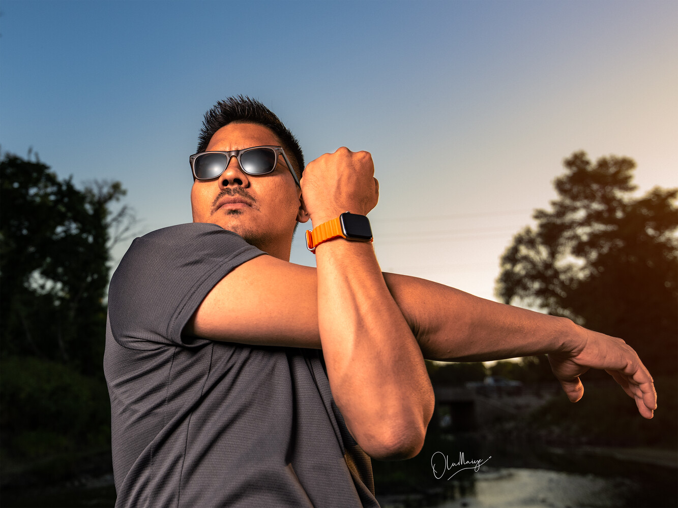 Man in sunglasses and blue shirt posing outdoors during golden hour with arms crossed
