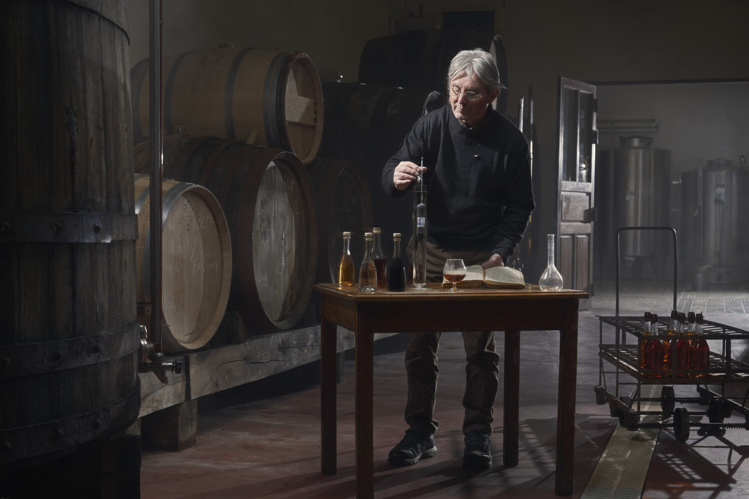 Older man with gray hair pouring amber liquid at a wooden table among oak barrels in a dim distillery cellar
