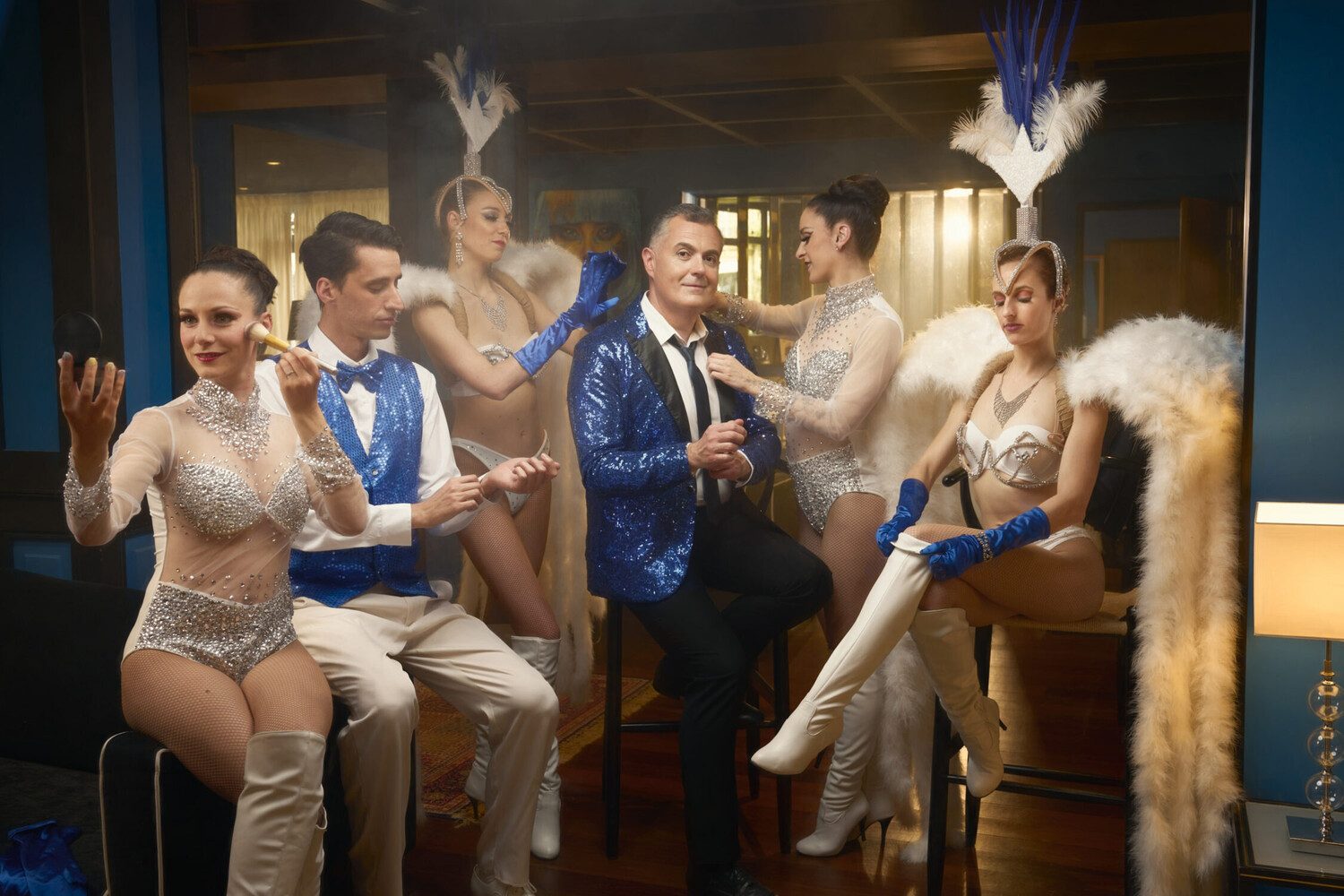 Man in a blue sequined jacket seated among four performers in sparkly showgirl costumes with white feathers and blue gloves in a warmly lit room.