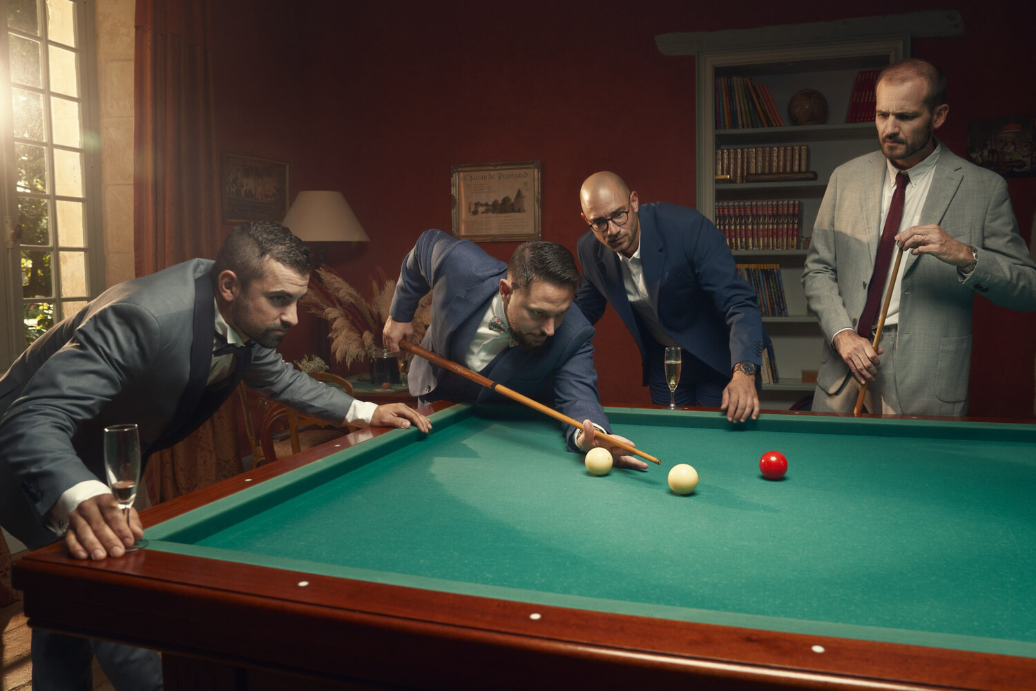 Four men in suits gather around a green felt pool table, one lining up a cue shot in a warm, wood-paneled room.