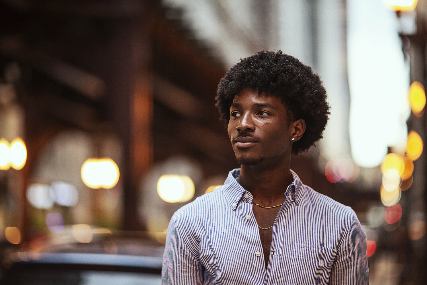 Young man in striped button-up shirt looking away, with blurred city street lights in background