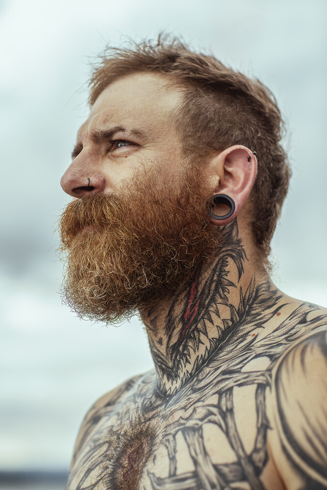 Man with full beard and neck tattoos in profile view against overcast sky