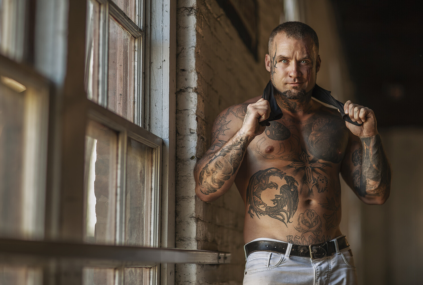 Shirtless man with extensive tattoos in boxing stance near industrial window
