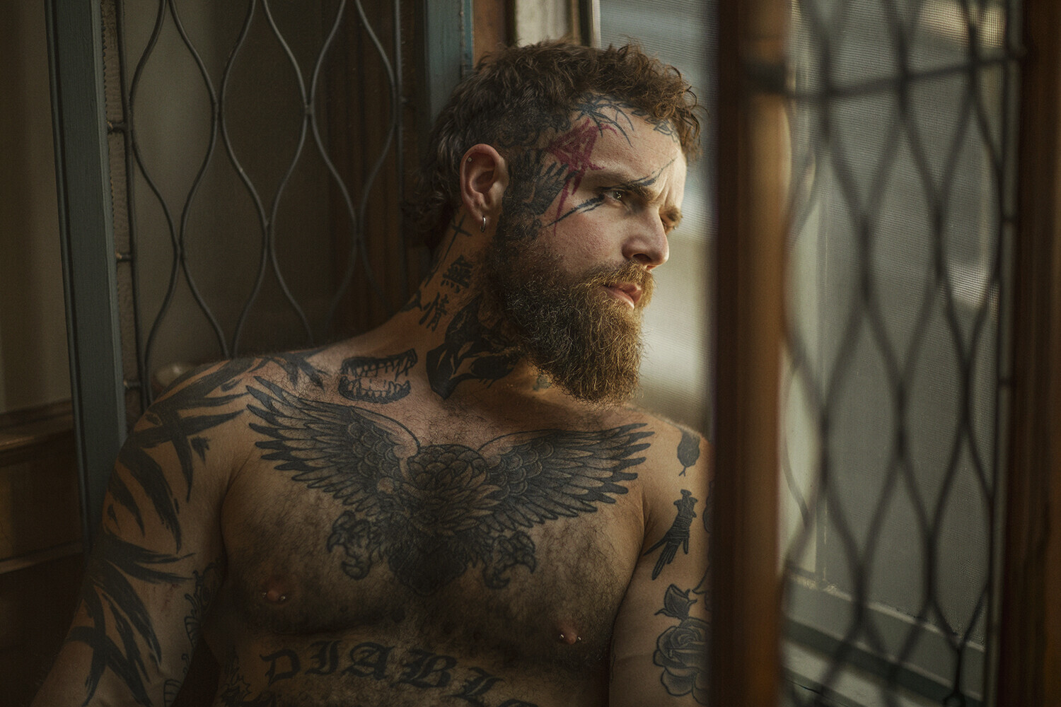 Shirtless man with extensive tattoos looking out through a chain-link window