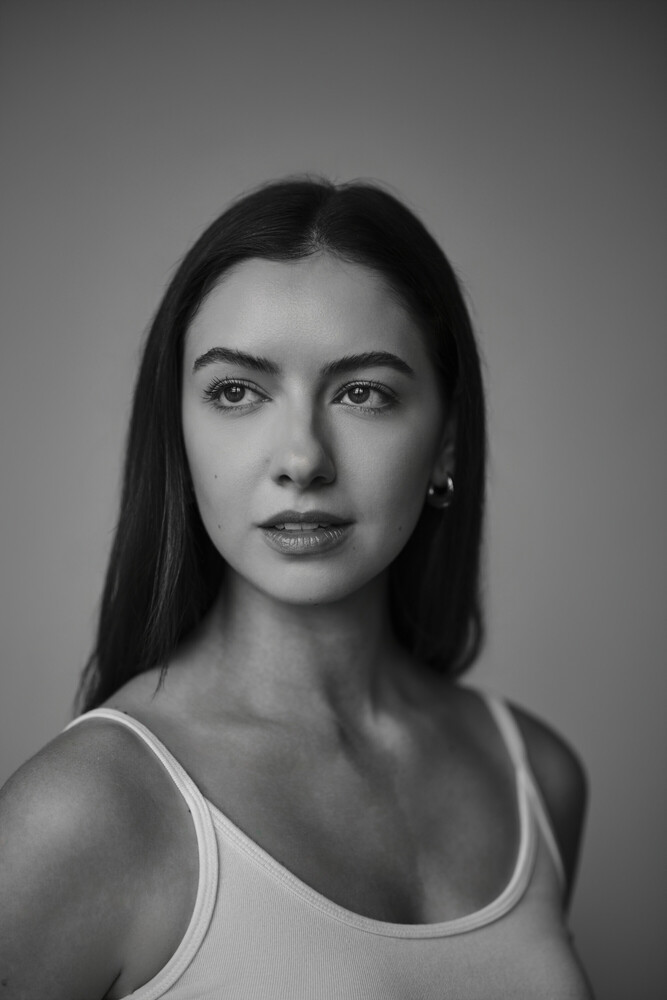 Black and white portrait of a woman with long dark hair wearing a white tank top against a neutral background