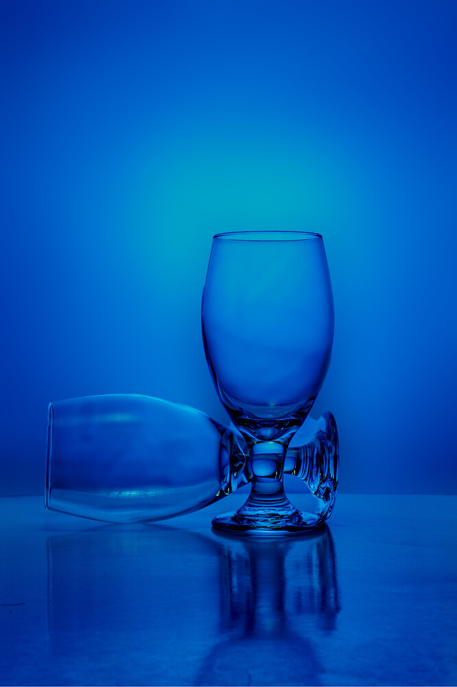 Empty and full drinking glasses on reflective surface against blue monochromatic background