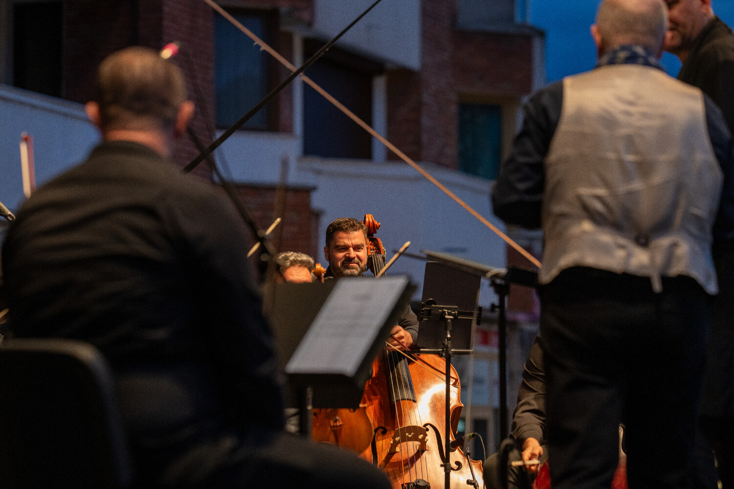 Orchestra conductor leading musicians during an outdoor evening performance with illuminated cello in foreground
