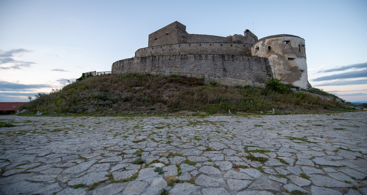 Stone fortress on grassy hilltop beside calm water at dusk