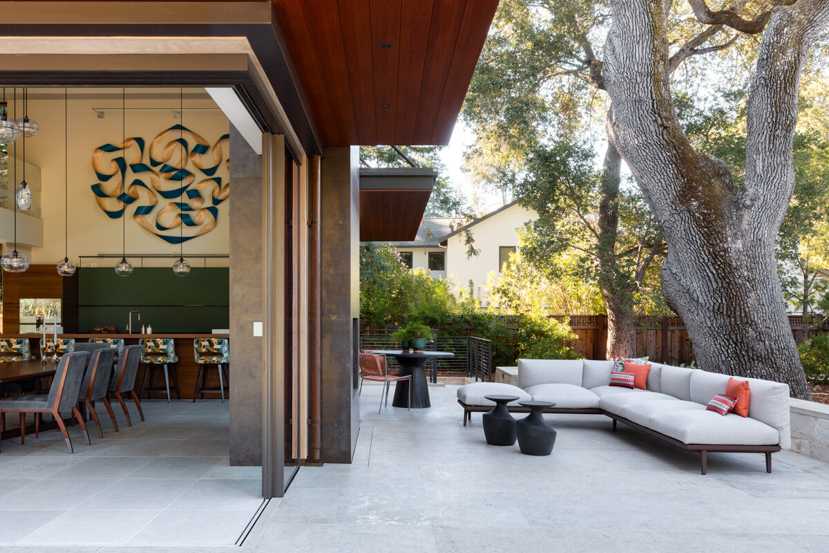 Covered patio with a white sectional sofa and orange pillows, open to a modern kitchen and dining area beside a large oak tree