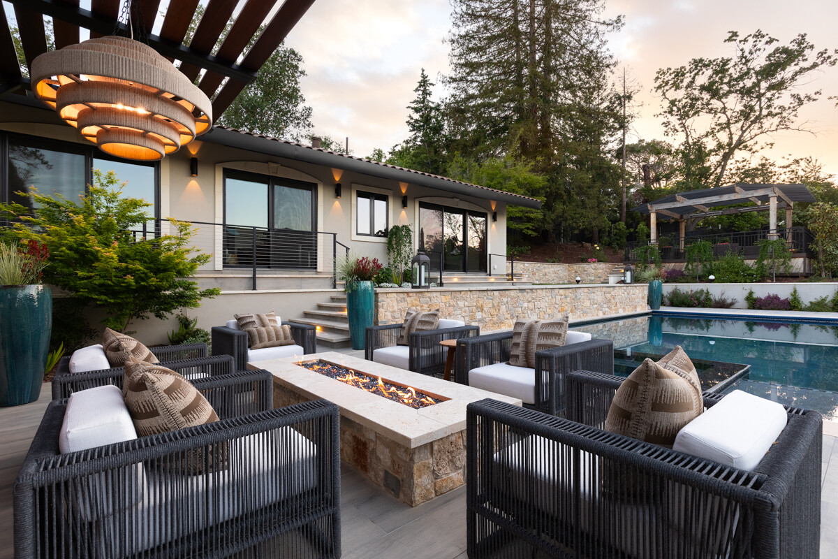Outdoor patio at dusk with a stone gas fire pit surrounded by black woven lounge chairs with white cushions, beside a lit pool and a large woven pendant lamp.