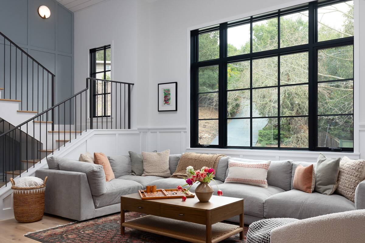 Modern living room with a light gray sectional sofa, wood coffee table, and a large black-framed window overlooking trees, beside a staircase with black railings.