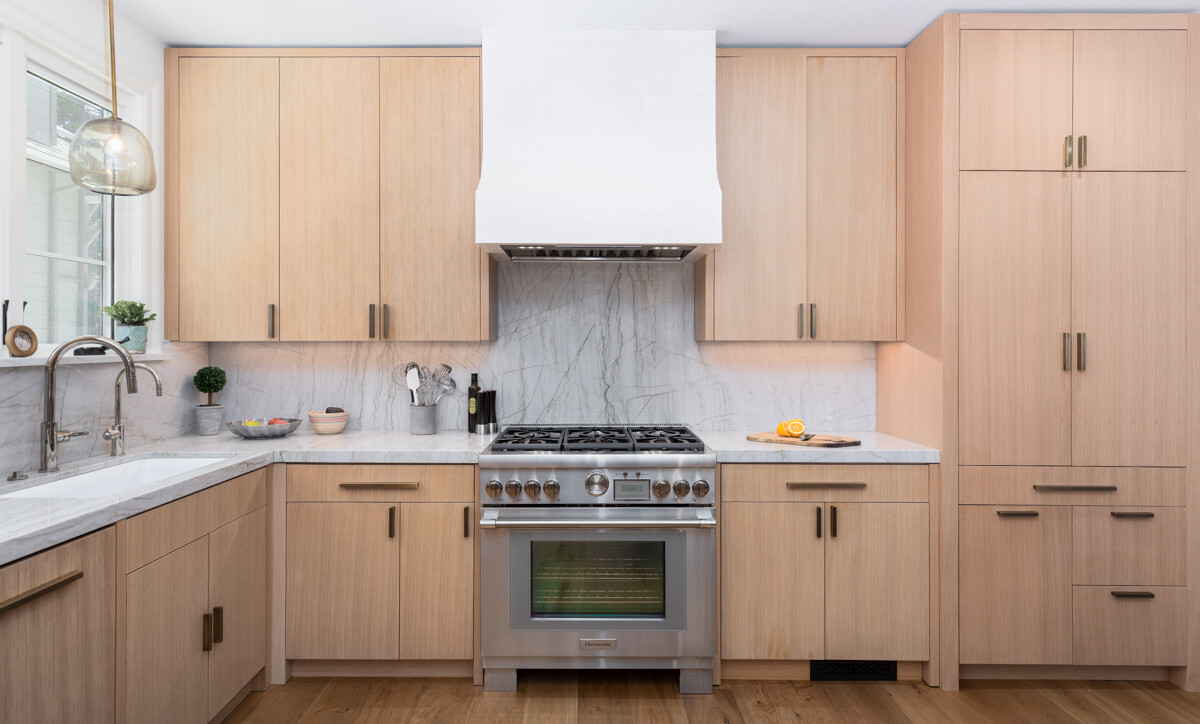 Modern kitchen with light oak flat-panel cabinets, white range hood, and marble backsplash beside a stainless steel range.
