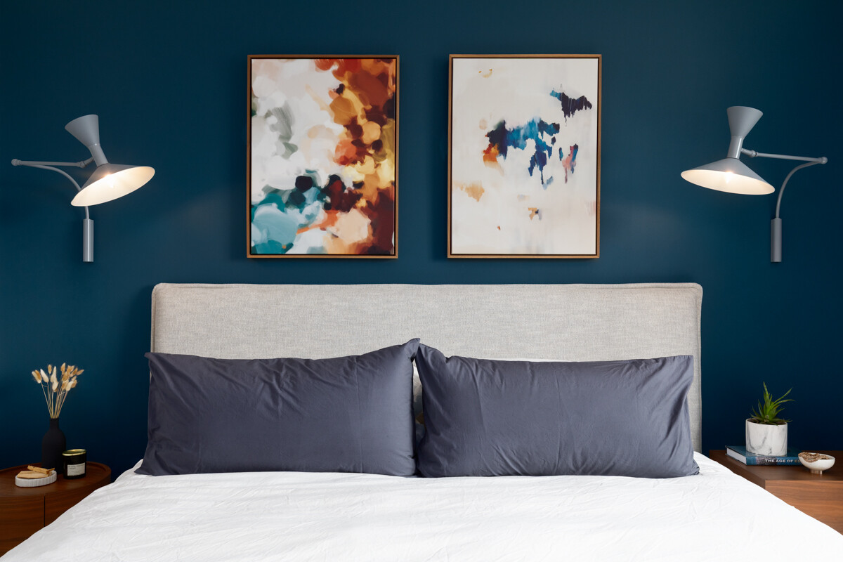 Bedroom with deep teal walls, a gray upholstered headboard, navy pillows, and two abstract paintings hanging above the bed.
