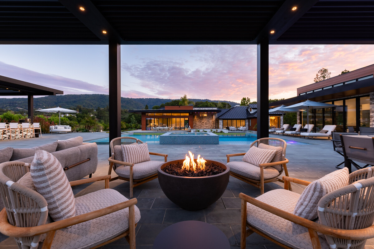 Outdoor patio at dusk with a lit round fire pit surrounded by cushioned wood-frame lounge chairs, with a pool and modern house in the background.