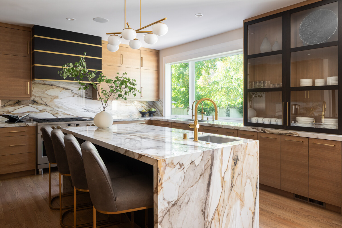 Modern kitchen with a waterfall-edge marble island with gold veining, brass faucet, and bar stools, beside a window with greenery outside.