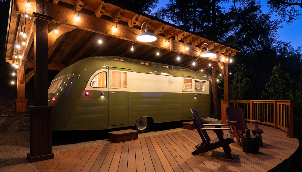 Vintage-style olive green and cream travel trailer on a wooden deck under a rustic pergola strung with warm lights at dusk.