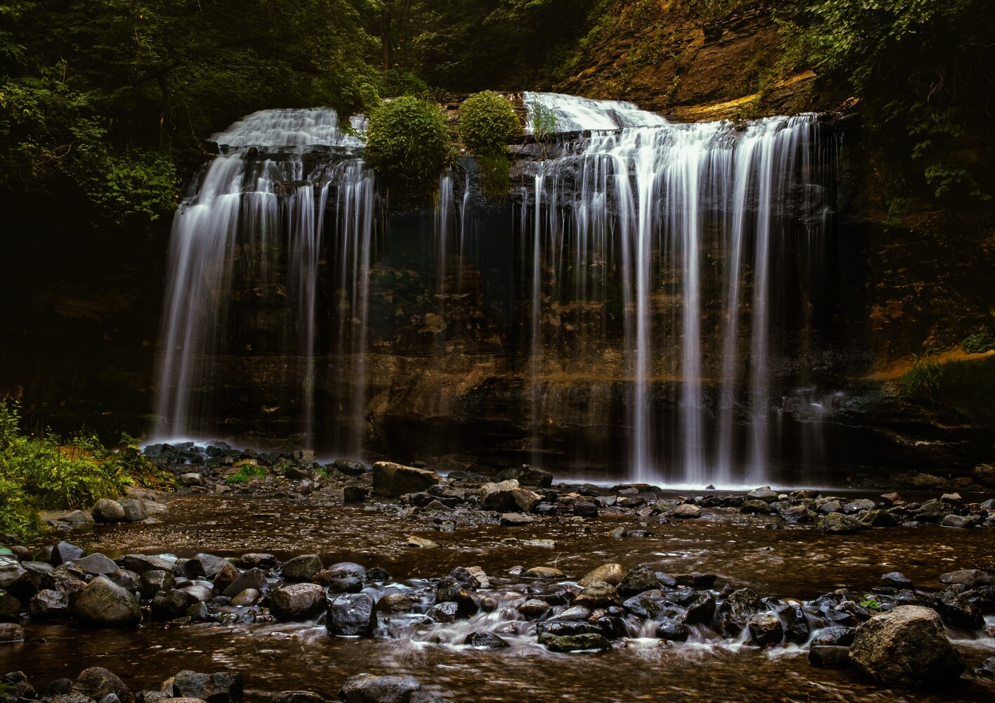 Cascading waterfall with silky water streams flowing over moss-covered rock ledge into shallow stream below
