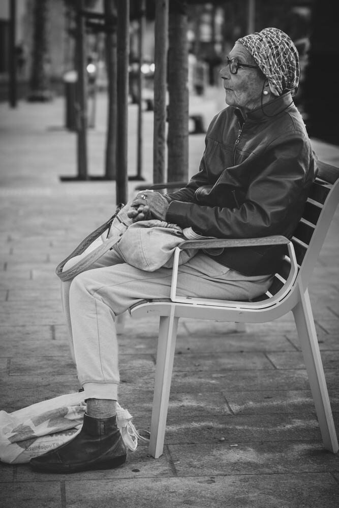 Person seated on a metal chair on an urban sidewalk, reading a newspaper