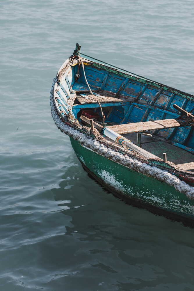 Weathered blue and green wooden fishing boat with rope and oars moored in calm water