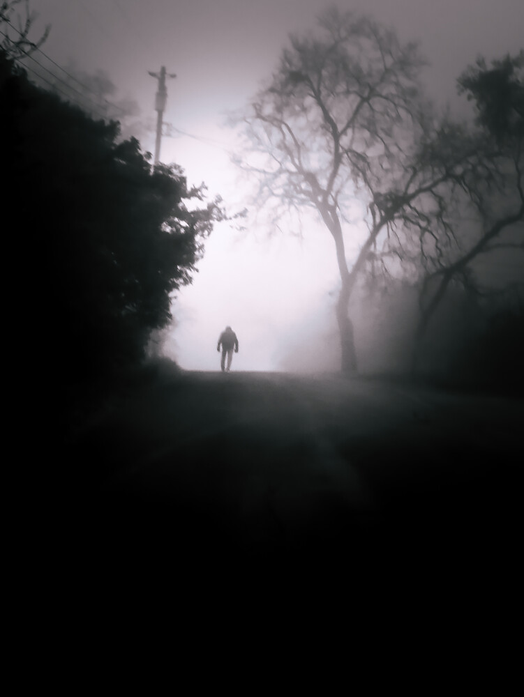 Solitary figure standing on a misty road beneath bare trees and a wooden cross