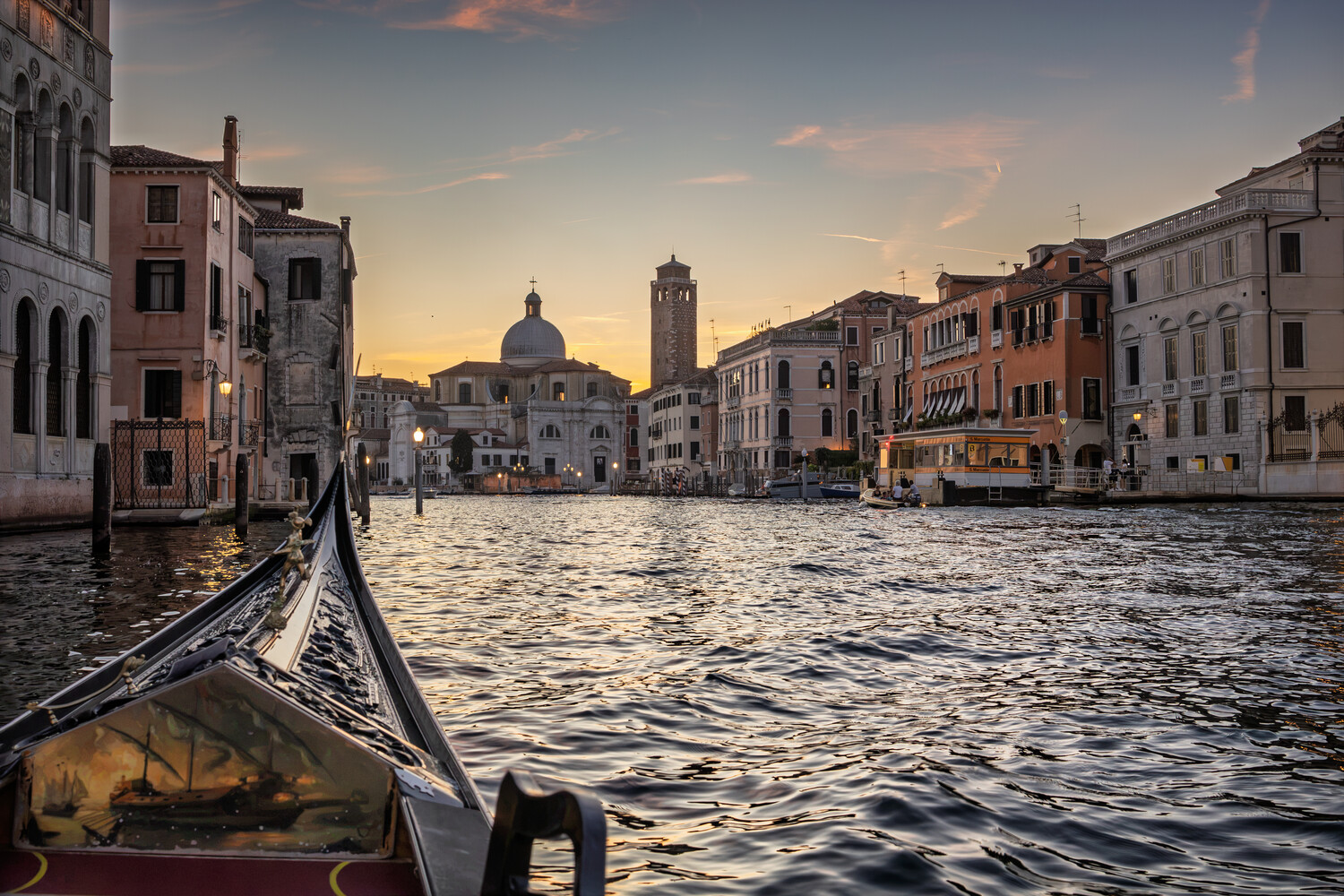 Gondola in foreground on Venice's Grand Canal at golden hour with basilica dome and bell tower in distance