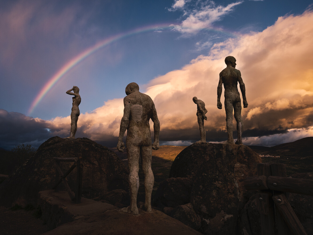 Five silhouetted figures standing on rocky outcrops beneath a dramatic sunset sky with a rainbow