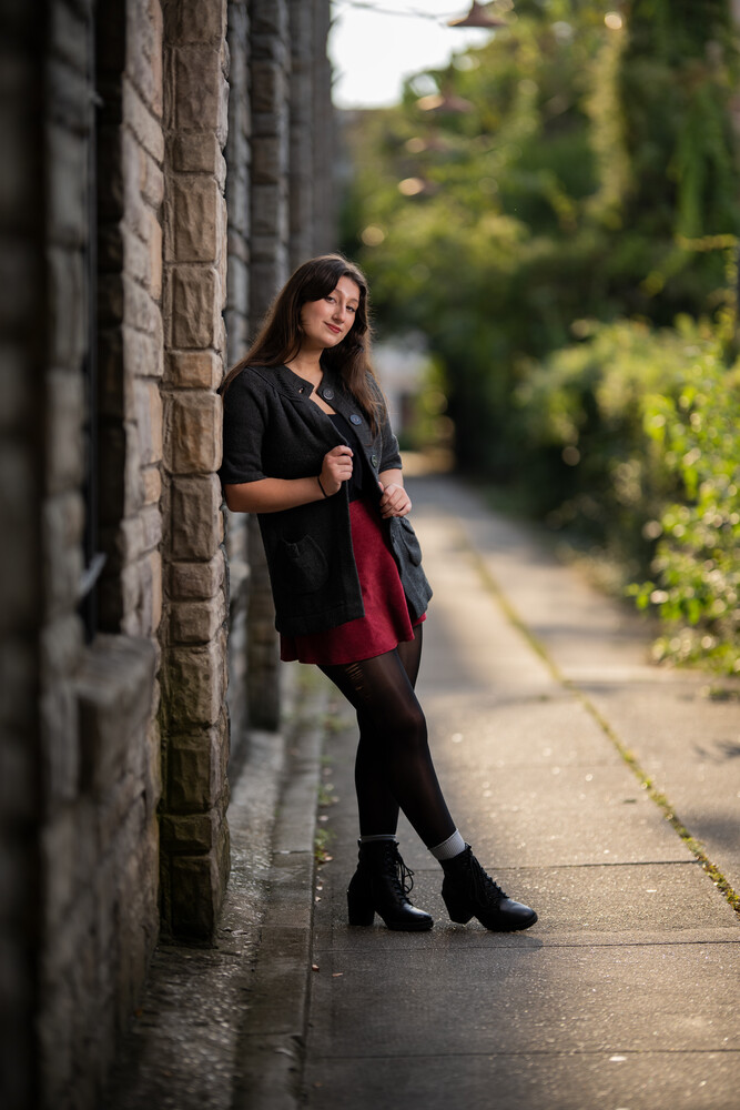 Woman in dark clothing leaning against brick wall on tree-lined urban pathway