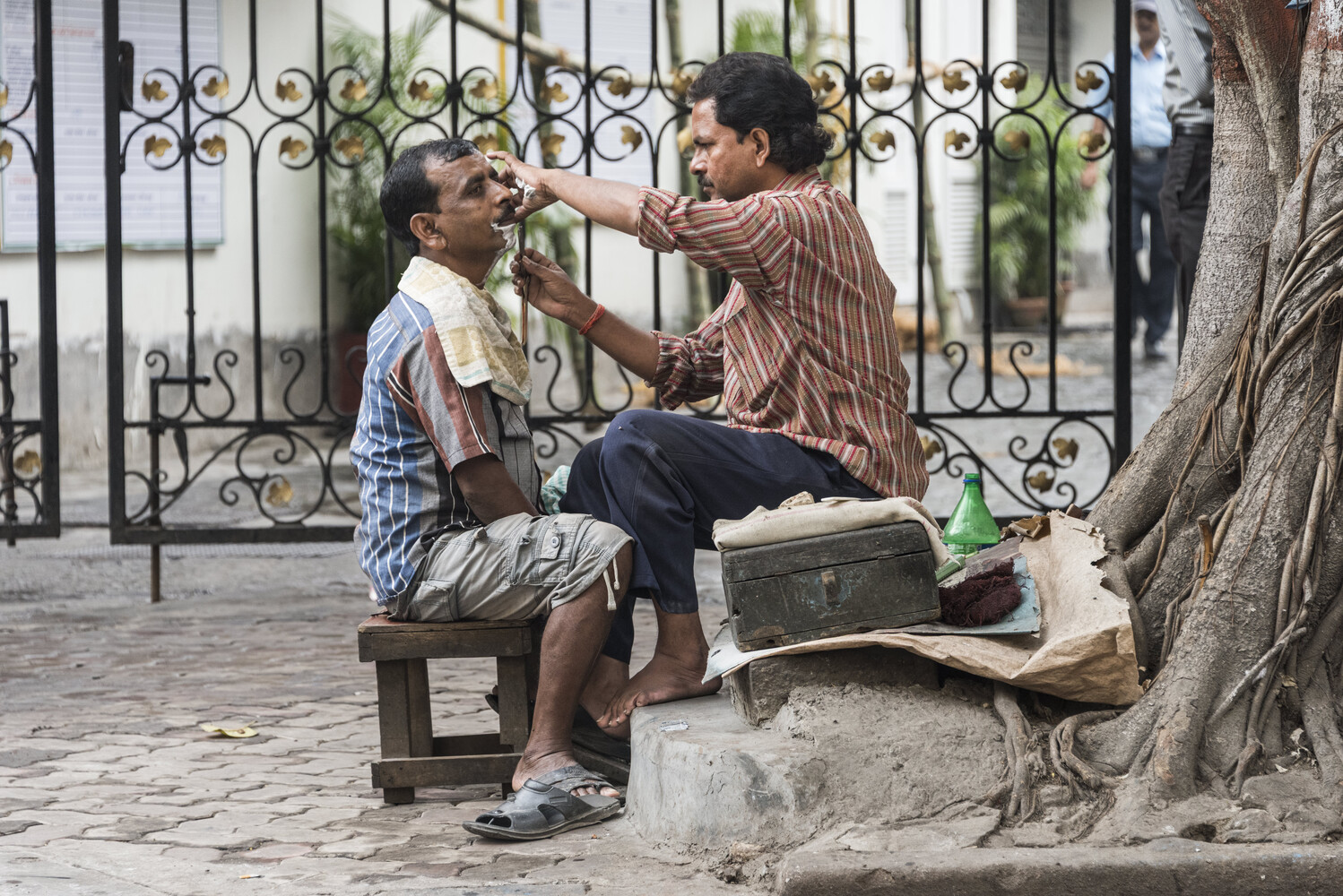 Man cutting another man's hair outdoors against ornamental iron gate backdrop