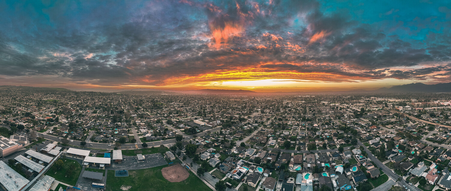 Aerial panorama of a sprawling suburban neighborhood at sunset beneath dramatic orange and teal clouds, with mountains on the horizon.
