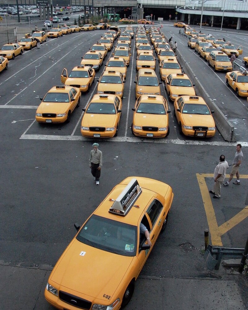 Yellow taxi cabs arranged in neat rows in a parking lot with pedestrians walking nearby
