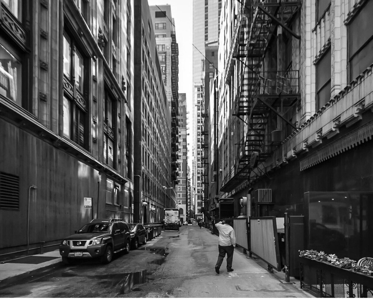 Black and white urban street scene with pedestrian walking between tall buildings and parked vehicles