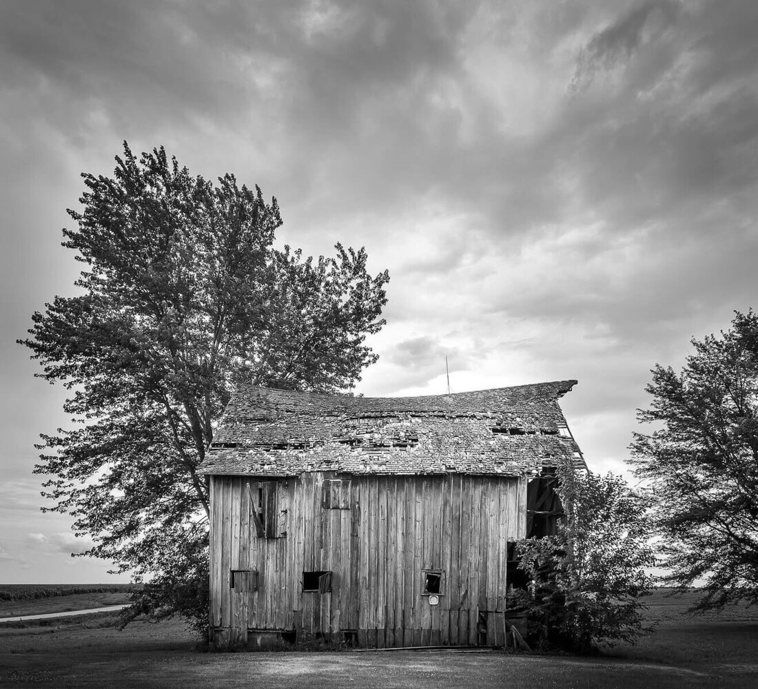 Abandoned wooden house with boarded windows, framed by windswept trees under stormy sky
