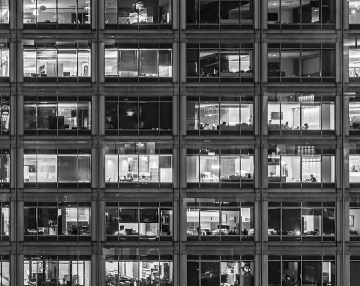 Black and white grid of office building windows showing interior workspaces at night