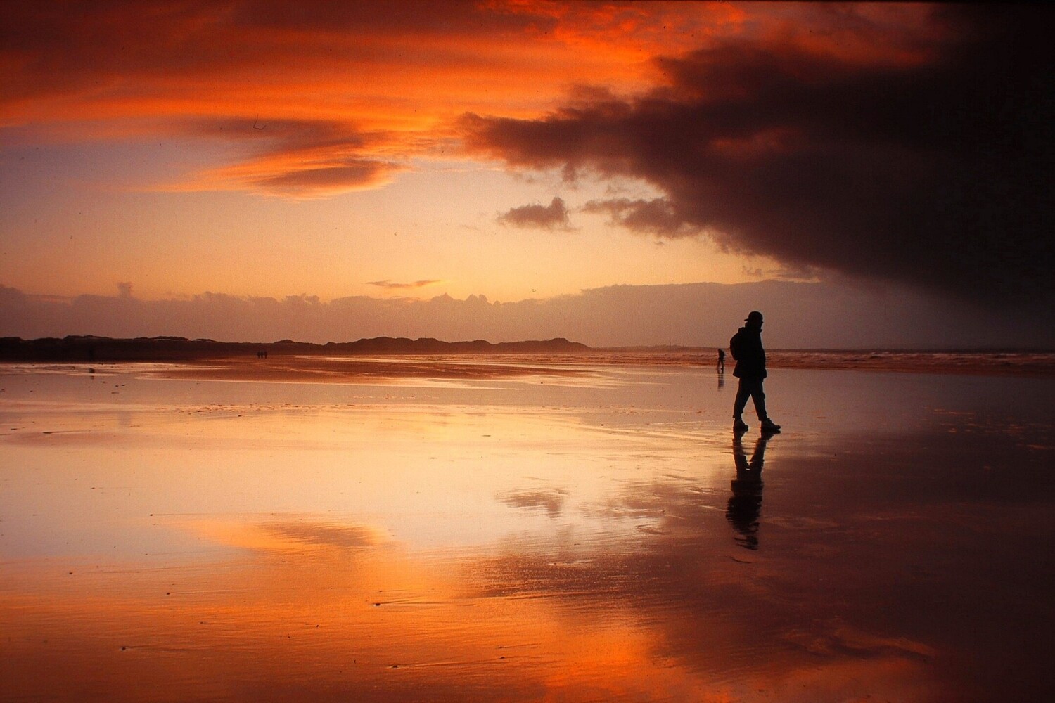 Silhouetted person walking across a wet beach at sunset, reflected in the shallow water under a fiery orange and dark cloud sky.