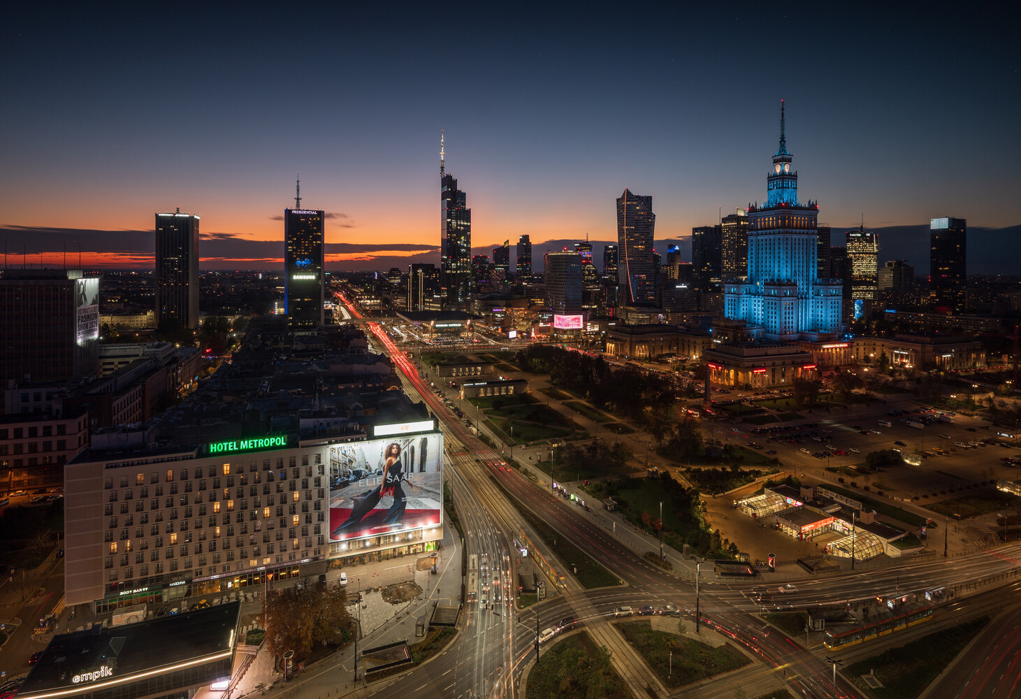 Aerial dusk view of a city skyline with the blue-lit Palace of Culture and Science and light trails on a busy boulevard