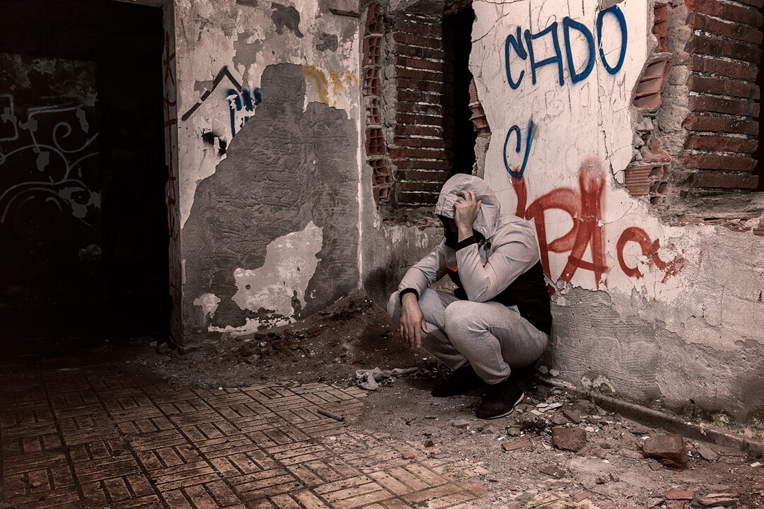 Person in light-colored hoodie crouching against weathered brick wall with graffiti