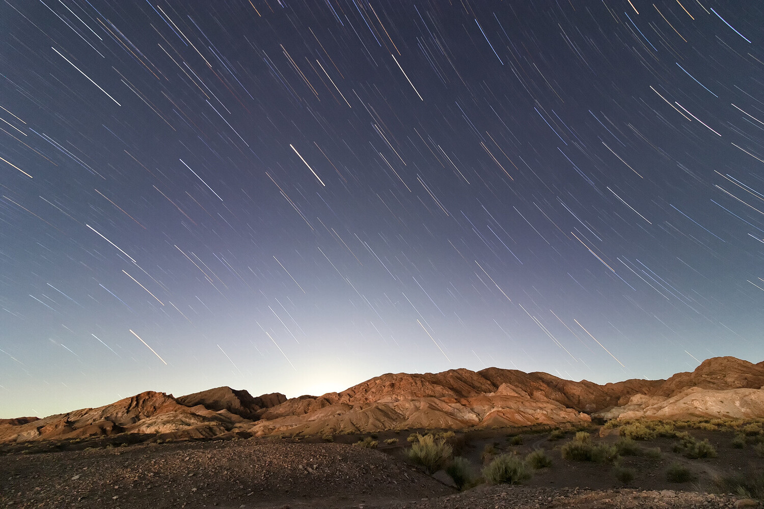 Desert landscape with star trails streaking across twilight sky above rocky mountains