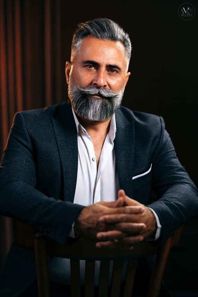 Bearded man in dark blazer and white shirt seated against dark backdrop