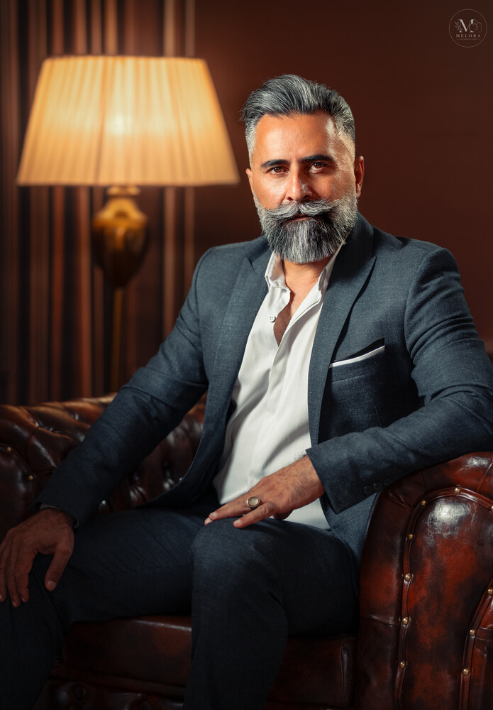 Bearded man in navy blazer and white shirt seated on leather chair against warm interior backdrop