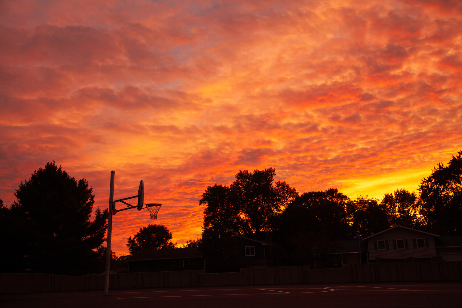 Basketball hoop silhouetted against dramatic fiery sunset sky with trees and houses