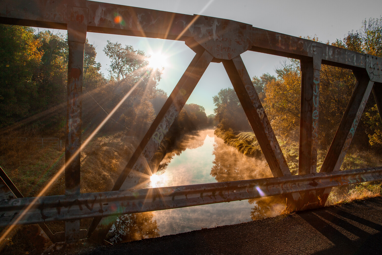 Rusty steel bridge frame overlooking a sunlit river lined with golden autumn foliage